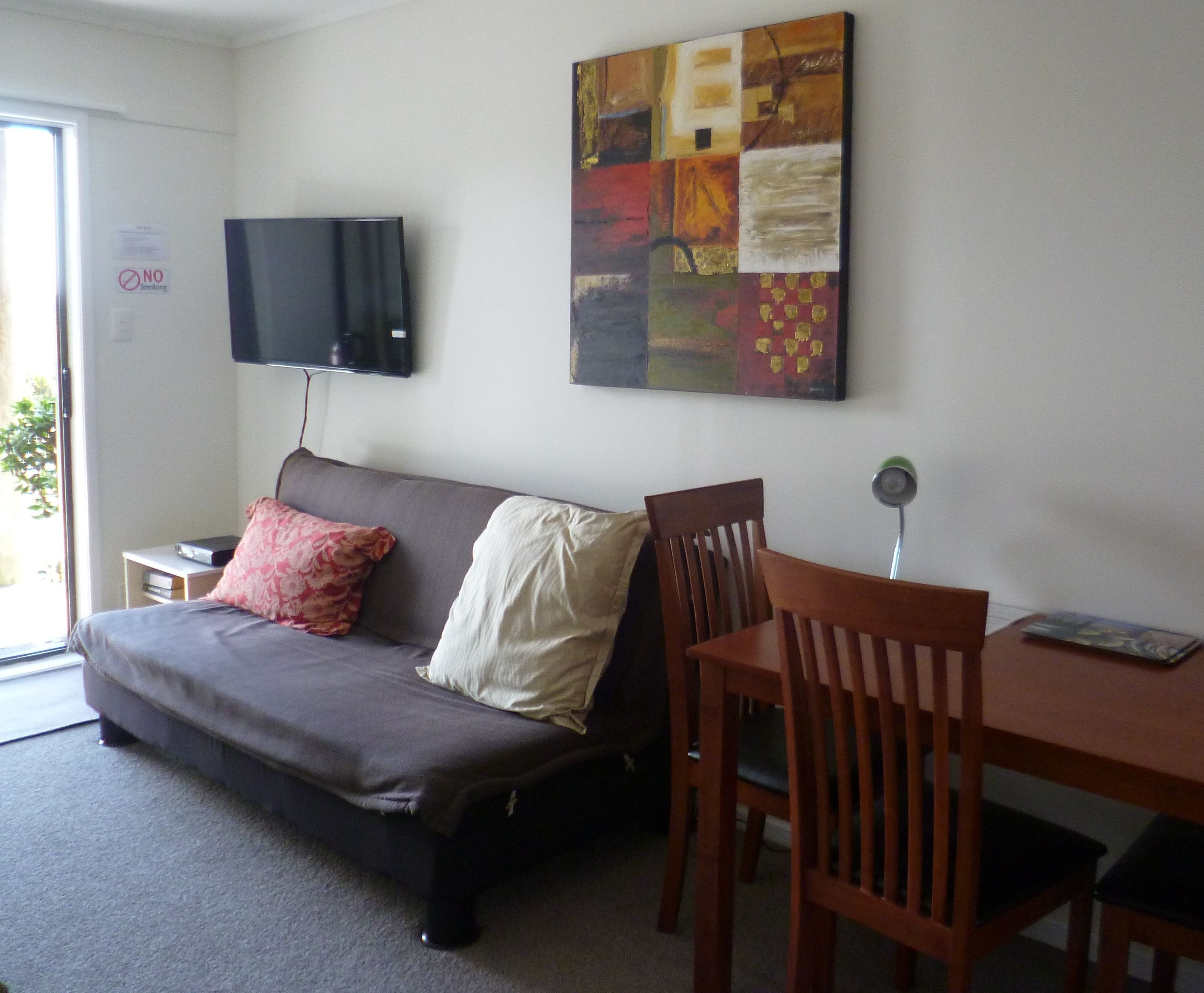 A room with a sofa, a TV mounted on the wall, a painting, a dining table with chairs, and a sign indicating no smoking.