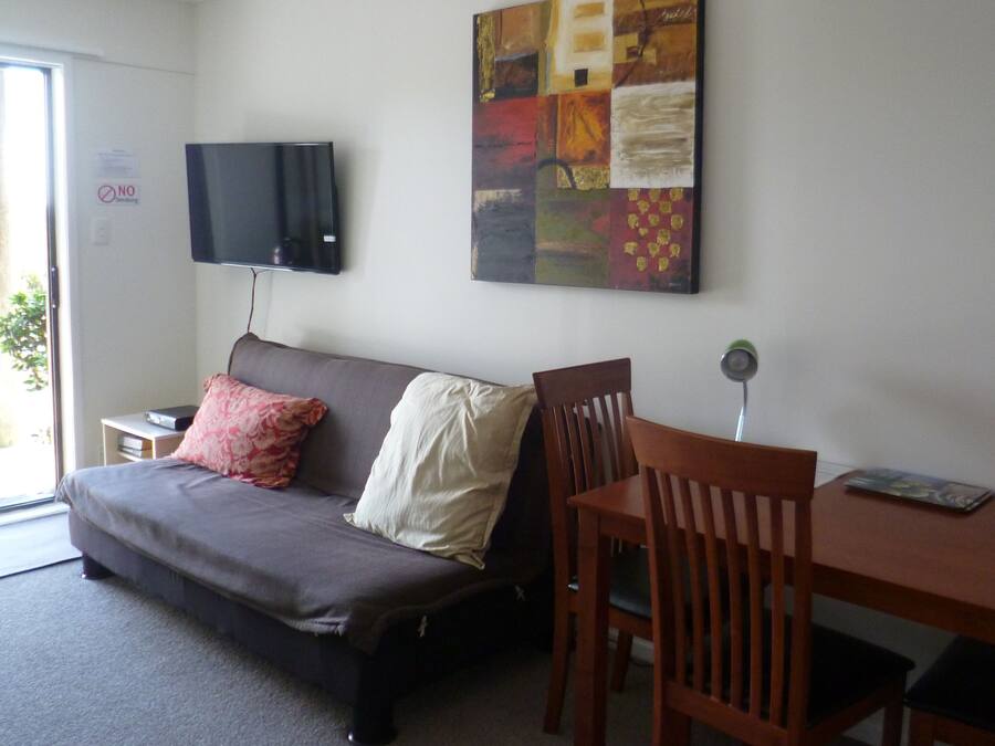 A room with a sofa, a TV mounted on the wall, a painting, a dining table with chairs, and a sign indicating no smoking.
