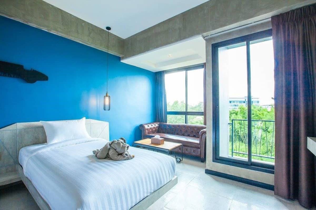 A modern hotel room with a large bed, a sofa, a small table, and a balcony with a view of greenery.