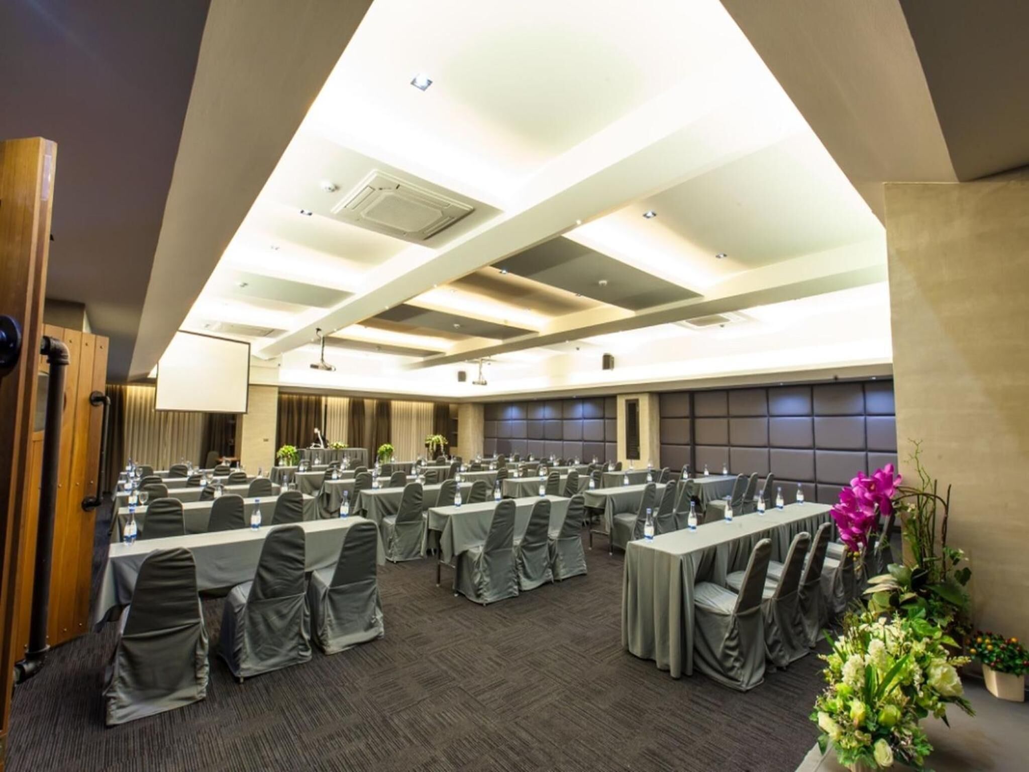 A conference room with multiple tables and chairs, equipped with water bottles and microphones, and a large screen at the front.