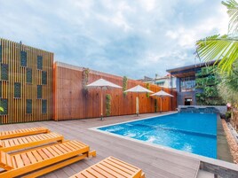 Una piscina con tumbonas de madera y sombrillas, rodeada por una valla de madera y un edificio moderno con balcón.
