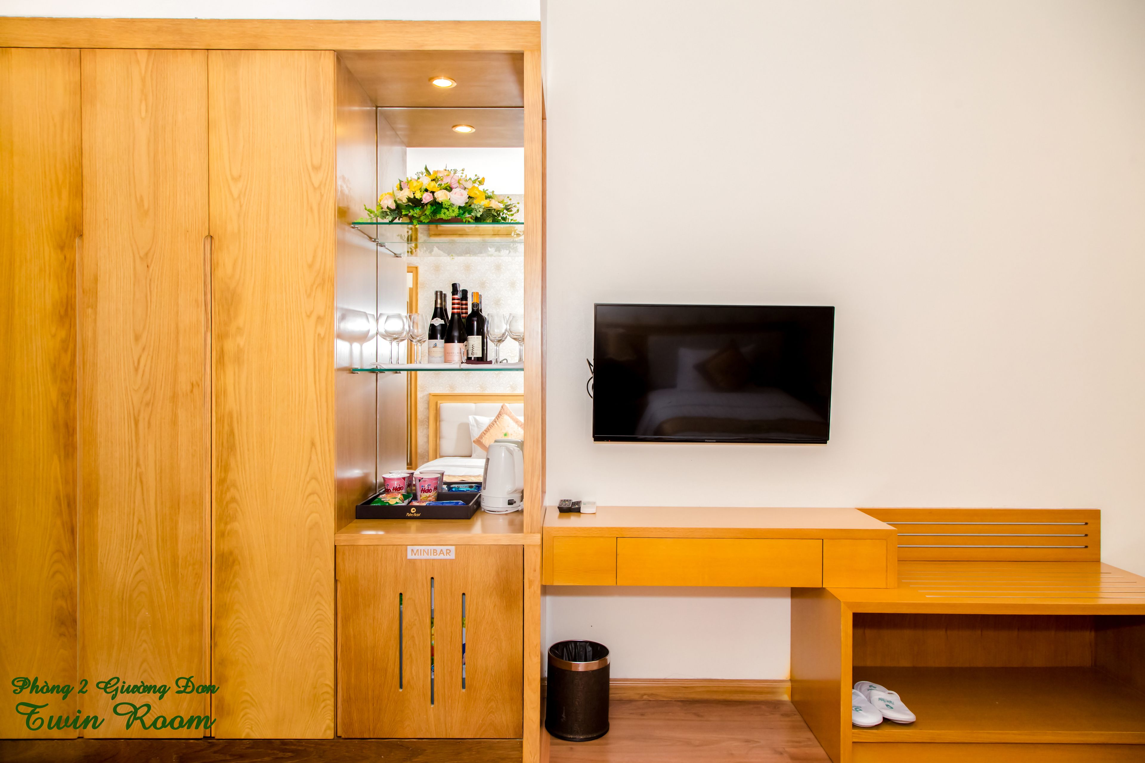 A twin room with a wooden wardrobe, a minibar, a TV, and a small seating area.