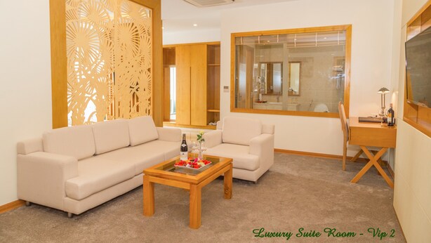 A luxury suite room with a beige sofa, a wooden coffee table with a tray of snacks, a wooden desk, and a large window. - Palm Hotel Thanh Hoa (Hac Thanh)