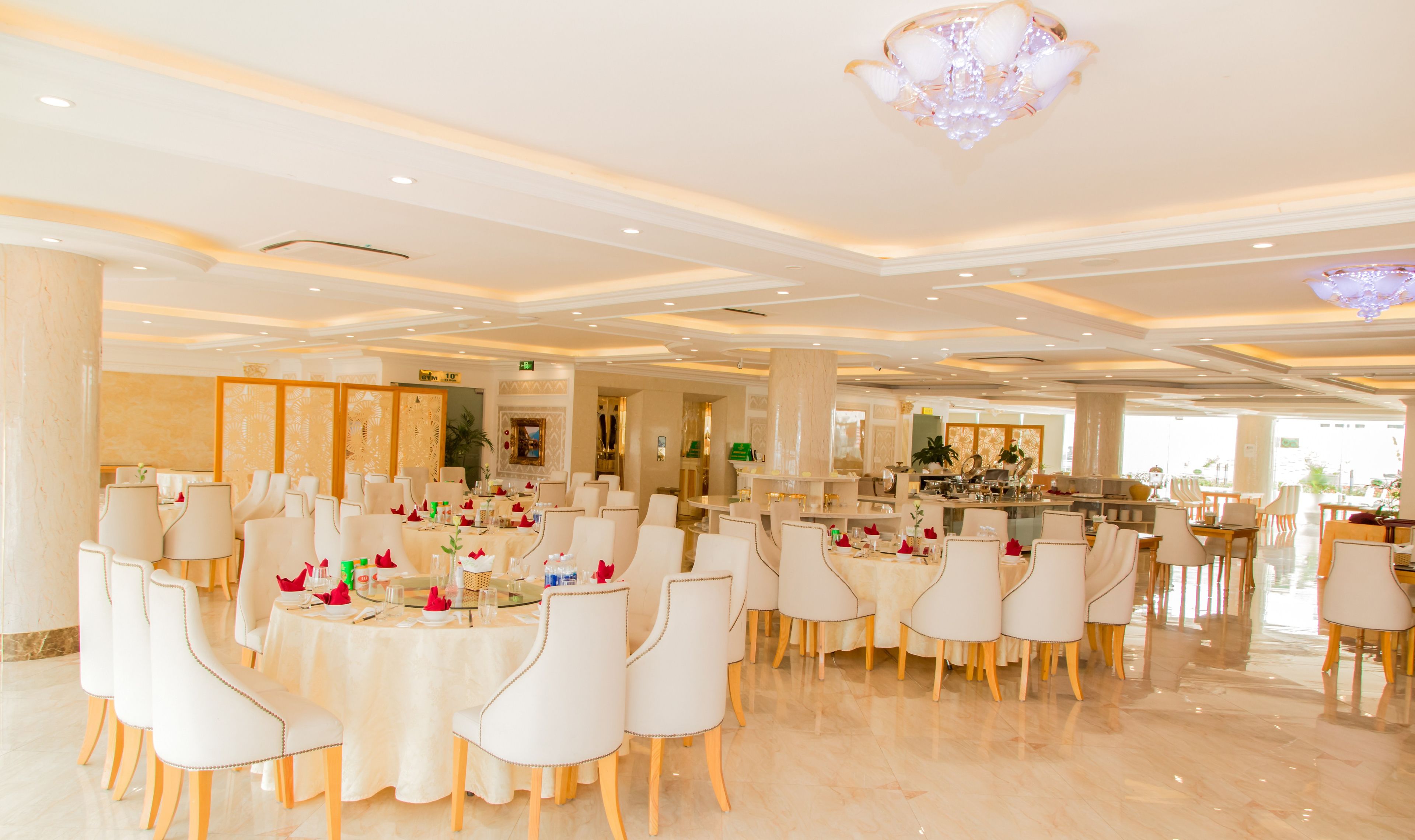 A spacious dining area with round tables and white chairs, featuring a chandelier and a view of the kitchen in the background.