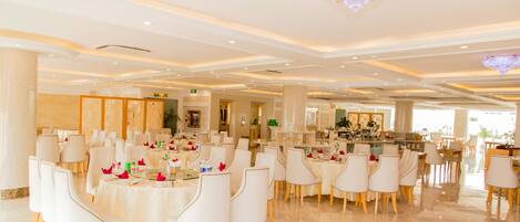 A spacious dining area with round tables and white chairs, featuring a chandelier and a view of the kitchen in the background.