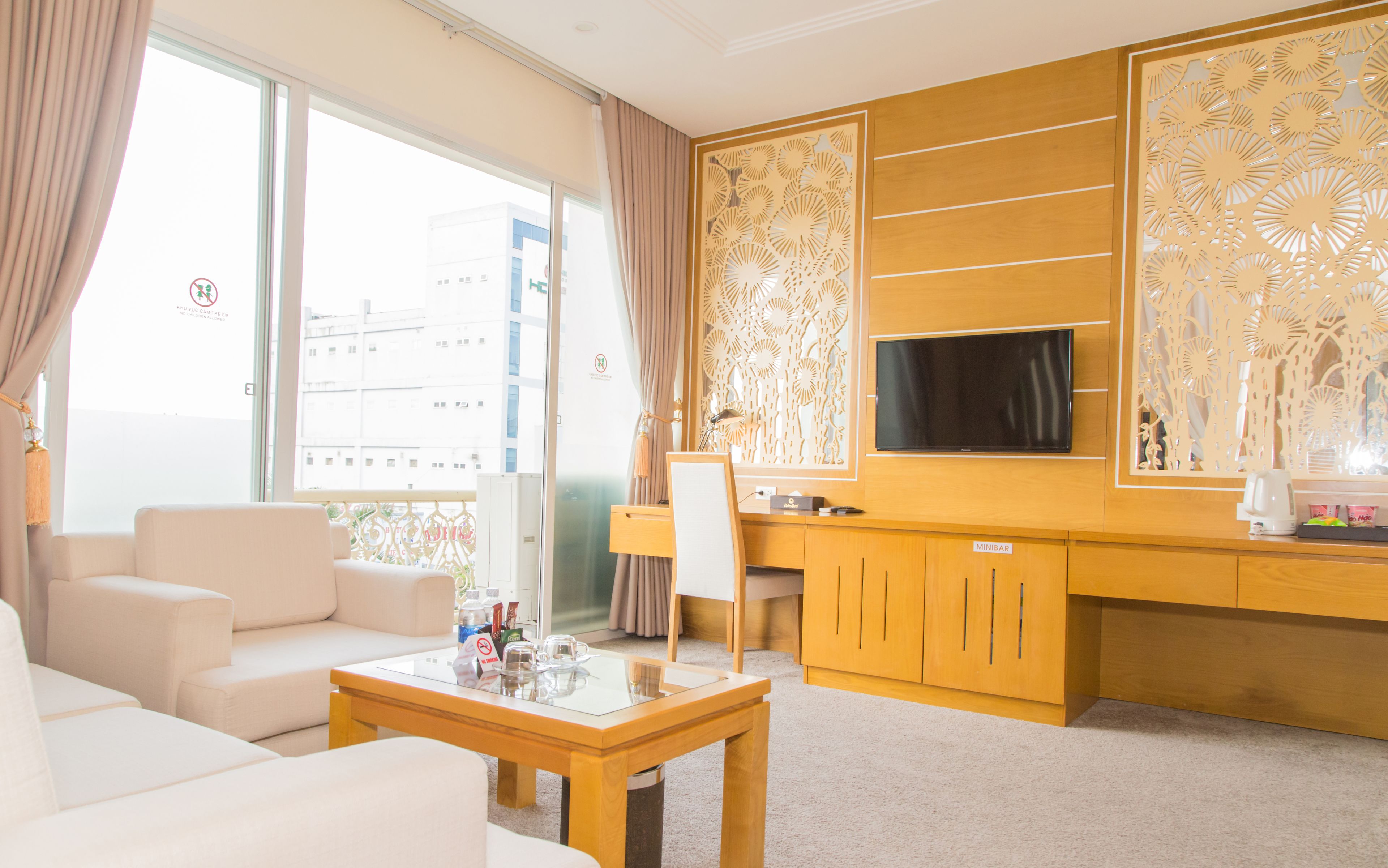A modern hotel room with a sofa, a glass-top coffee table, a desk with a chair, a television mounted on the wall, and a decorative wooden panel.