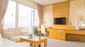 A modern hotel room with a sofa, a glass-top coffee table, a desk with a chair, a television mounted on the wall, and a decorative wooden panel.
