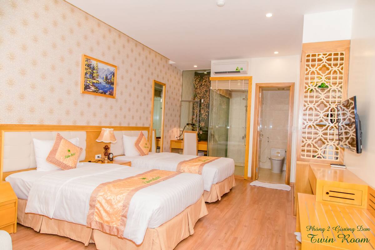 Een tweepersoonskamer met twee bedden, een houten bureau, een televisie en een badkamer met een glazen deur.