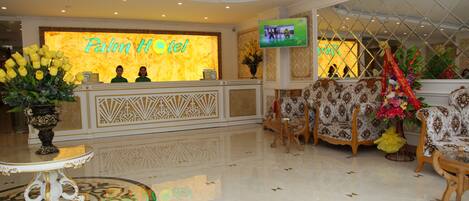 Zona de recepción de hotel con un mostrador, una pantalla de televisión y un área de descanso con sofás estampados.