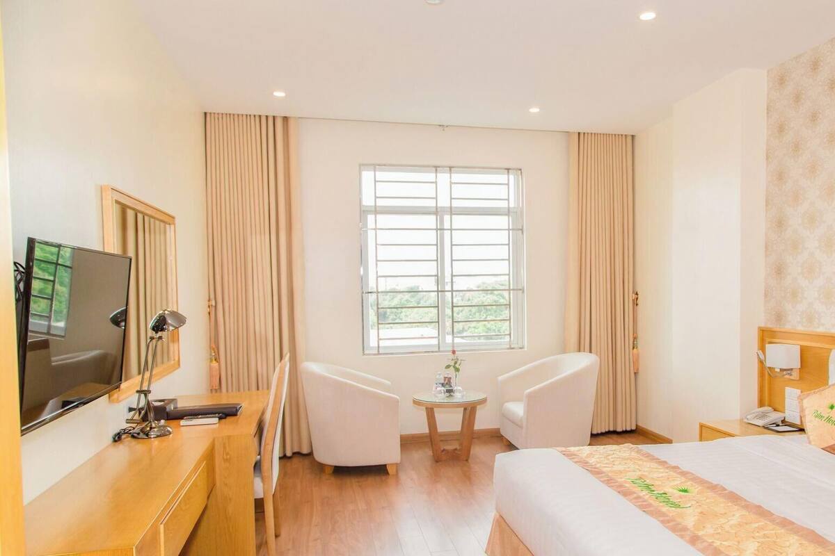 Een hotelkamer met een bed, een bureau met een computer, twee stoelen, een raam met gordijnen en een schilderij aan de muur.