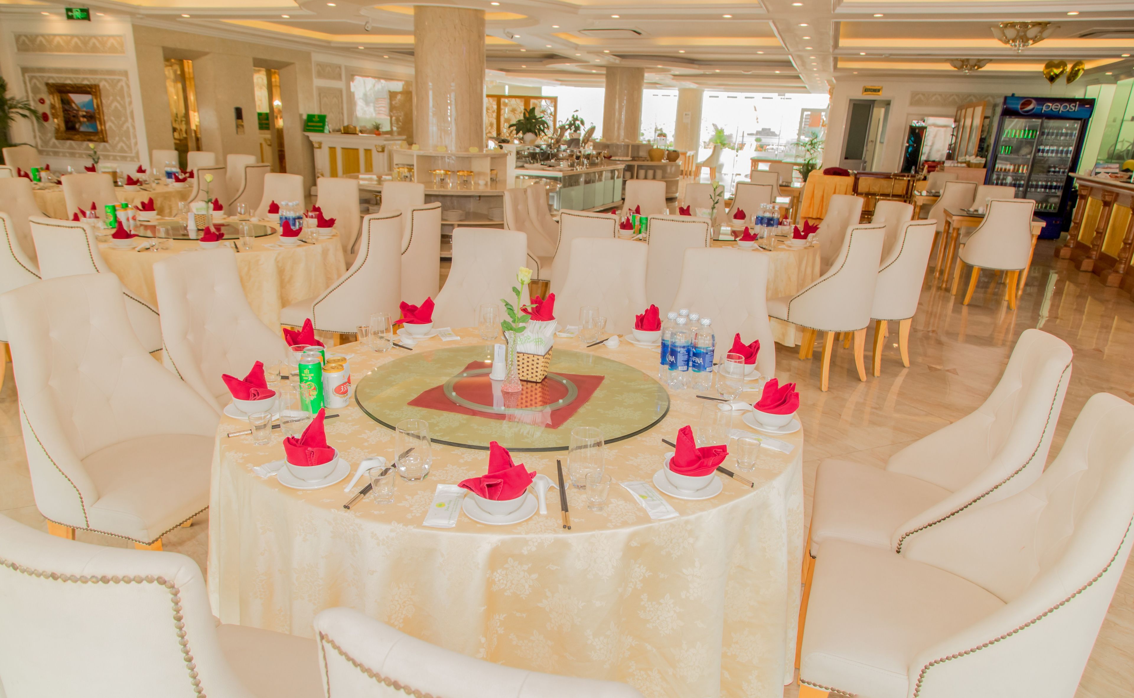 A dining area with round tables, white chairs, and a buffet counter in the background.