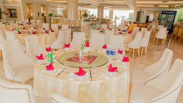 A dining area with round tables, white chairs, and a buffet counter in the background.