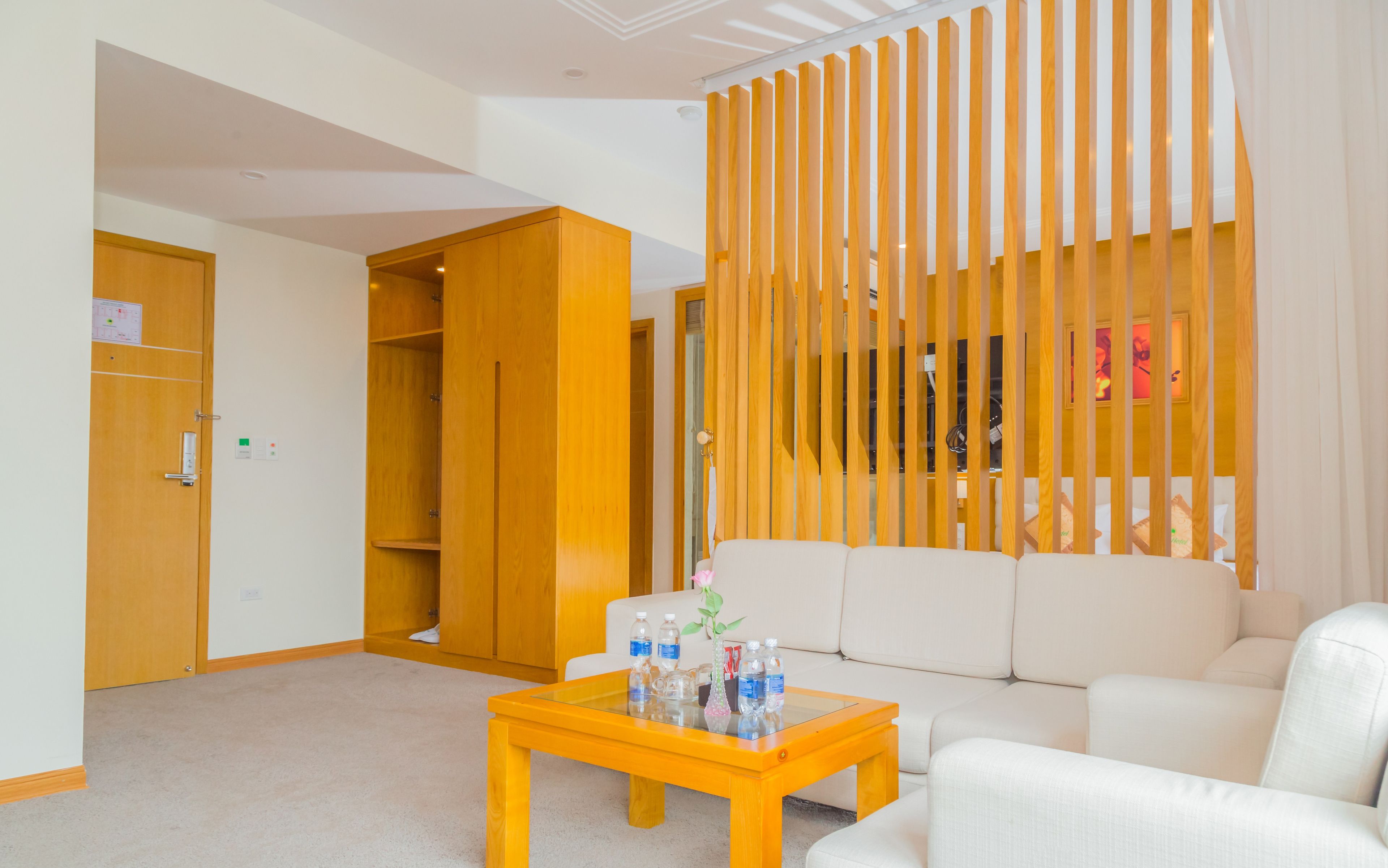 A hotel room with a wooden wardrobe, a white sofa, a glass-top coffee table with water bottles, and a wooden partition with vertical slats.