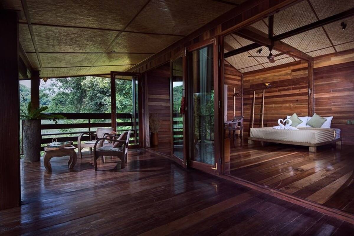 A wooden cabin with a large bed, a desk, and a chair. There is a balcony with a railing and a plant.