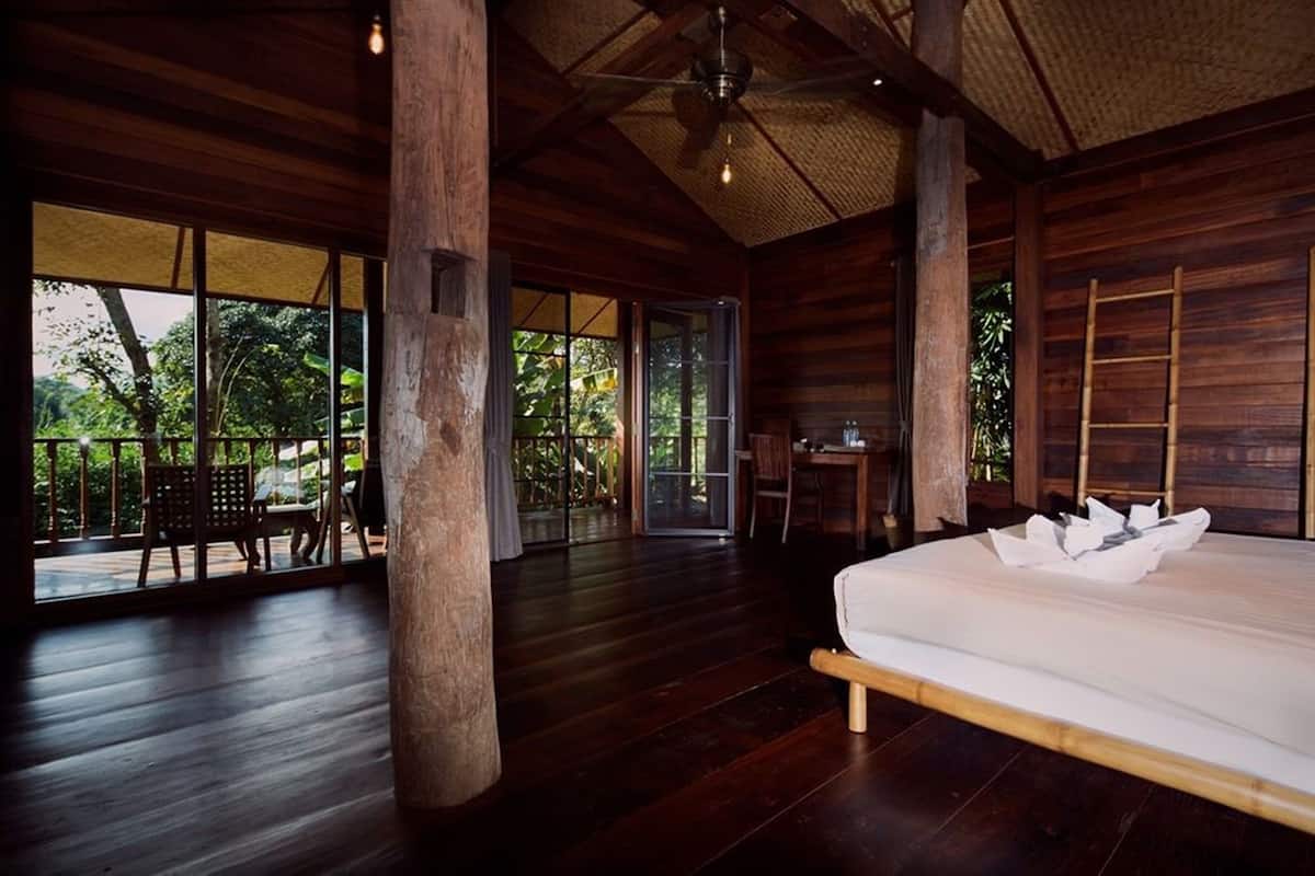 A spacious room with a large bed, wooden flooring, and a view of lush greenery outside.