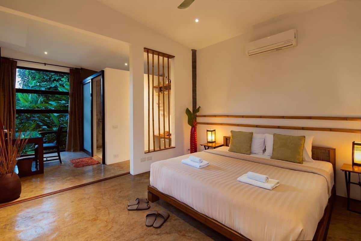 A modern hotel room with a large bed, a small table, a chair, and a view of greenery through a sliding door.