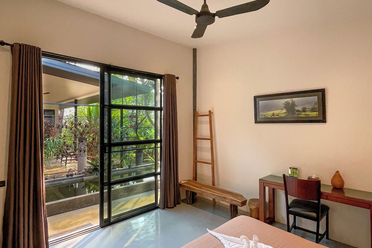 A hotel room with a bed, a desk, a chair, a ceiling fan, and a view of greenery outside.