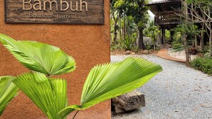 Ein hölzernes Schild mit dem Text "Bambuh Boutique Homestay" und Blick auf ein umgeben von Grünanlagen befindliches Gebäude.