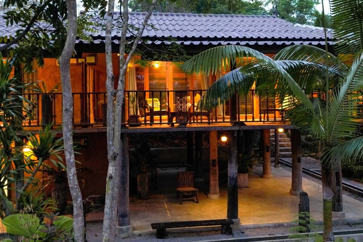A two-story building with a balcony, surrounded by tropical plants.