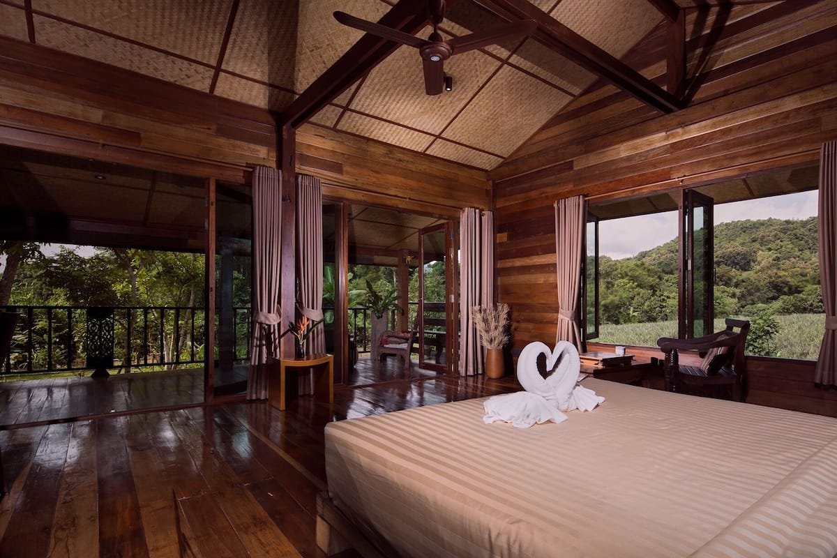 A spacious wooden room with a large bed, a dining table, and a view of a lush forest.