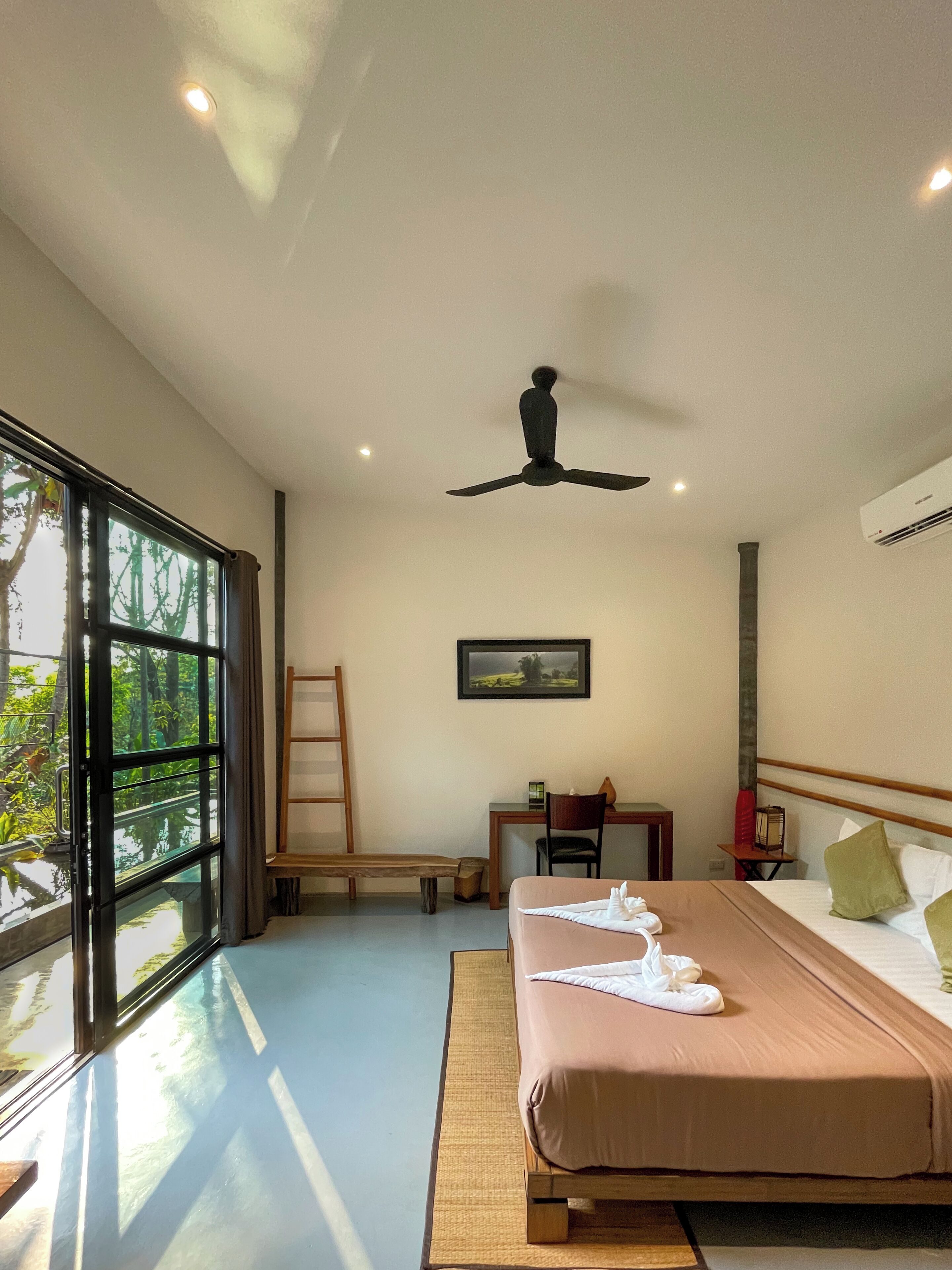 A hotel room with a bed, a desk, a chair, a ceiling fan, and a large window with a view of greenery.
