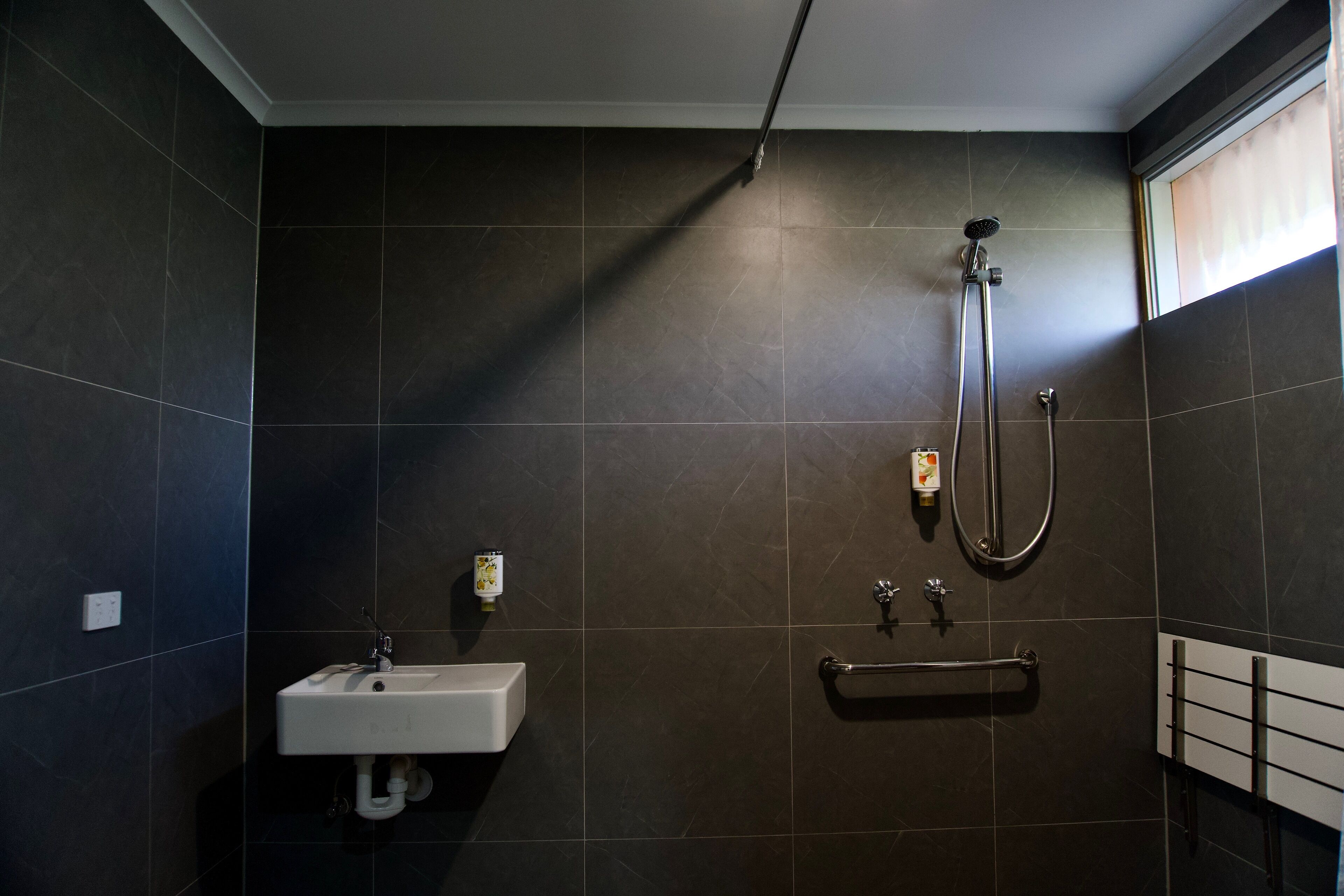 A bathroom with dark tiles, a white sink, a handheld showerhead, and a grab bar.