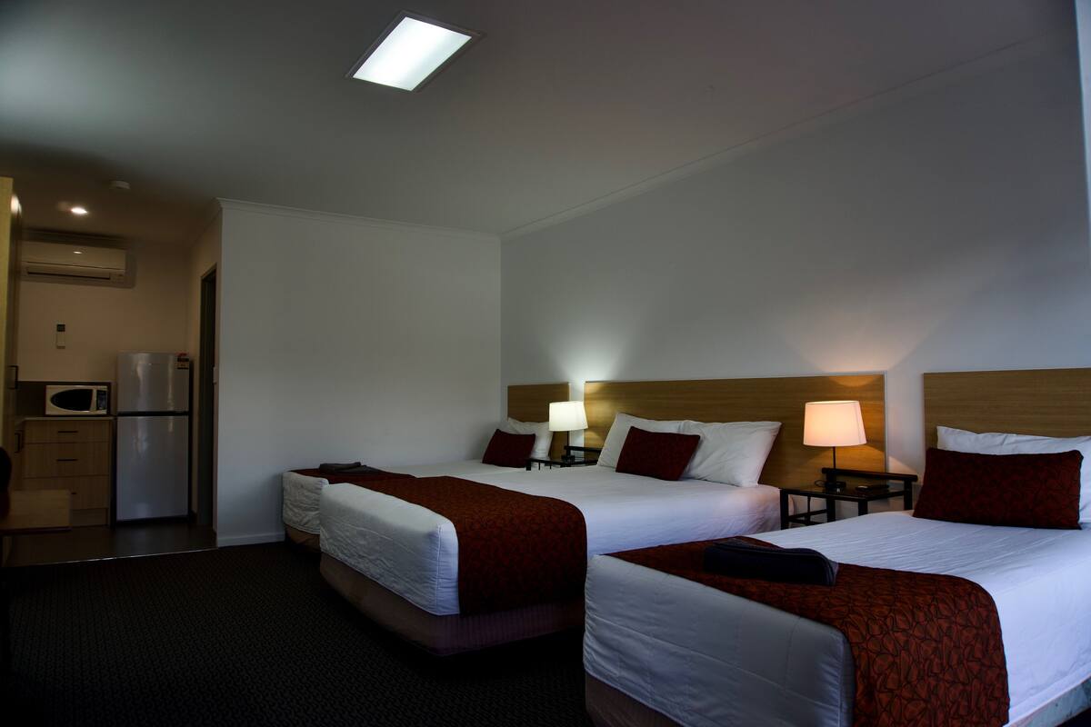 A hotel room with two beds, each with a red and white bedspread, a bedside table with a lamp, and a small desk with a refrigerator.
