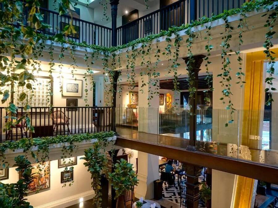 Un vestíbulo de hotel de varios niveles con barandilla de cristal, plantas de interior y obras de arte en las paredes.
