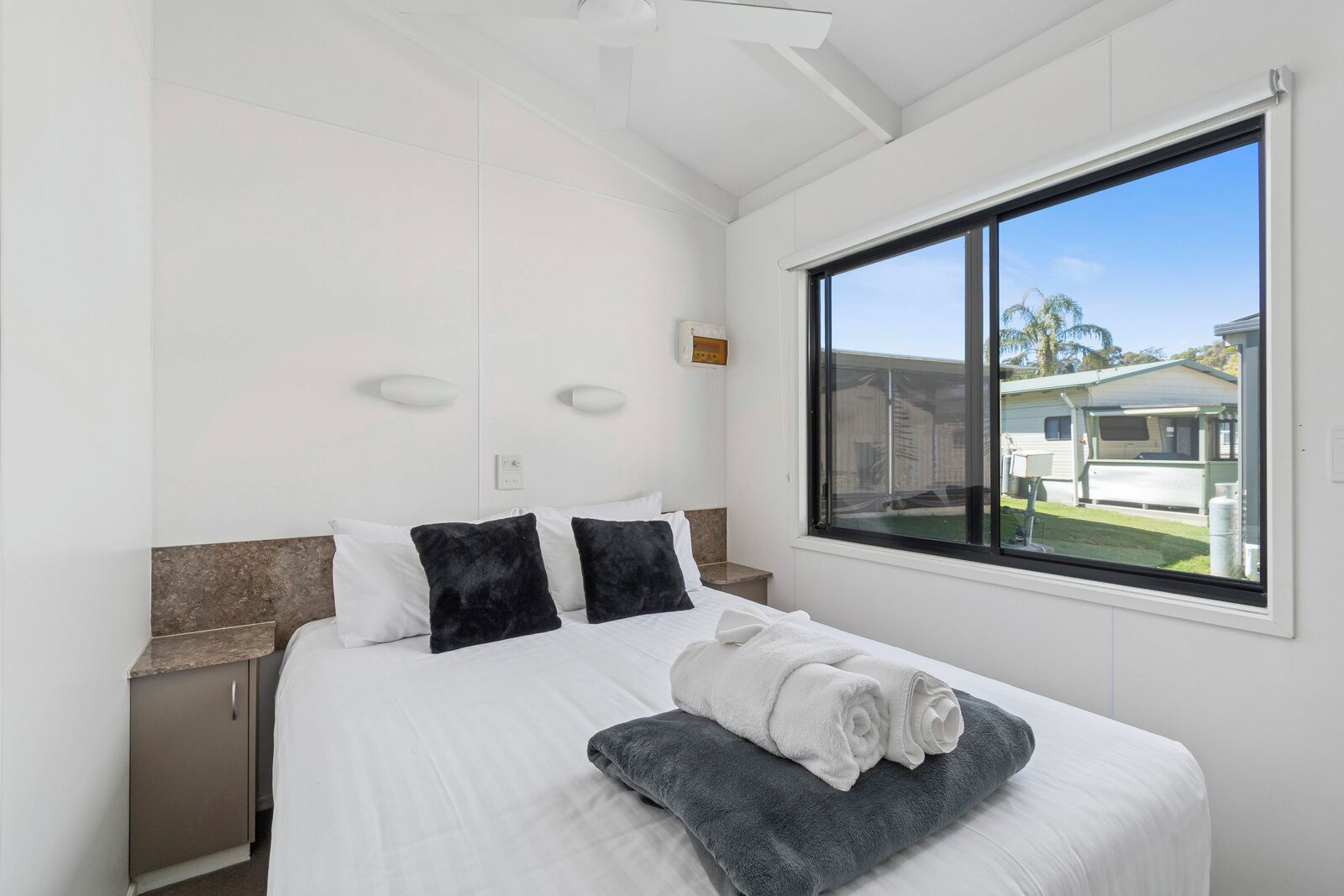 A bedroom with a bed, two bedside tables, a window with a view of houses, and a ceiling fan.