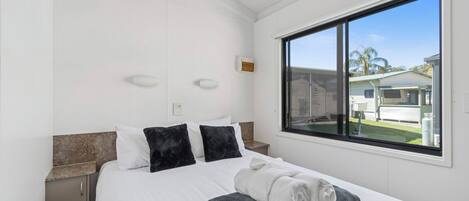 Quarto com cama, duas mesas de cabeceira, janela com vista para casas e ventilador de teto.