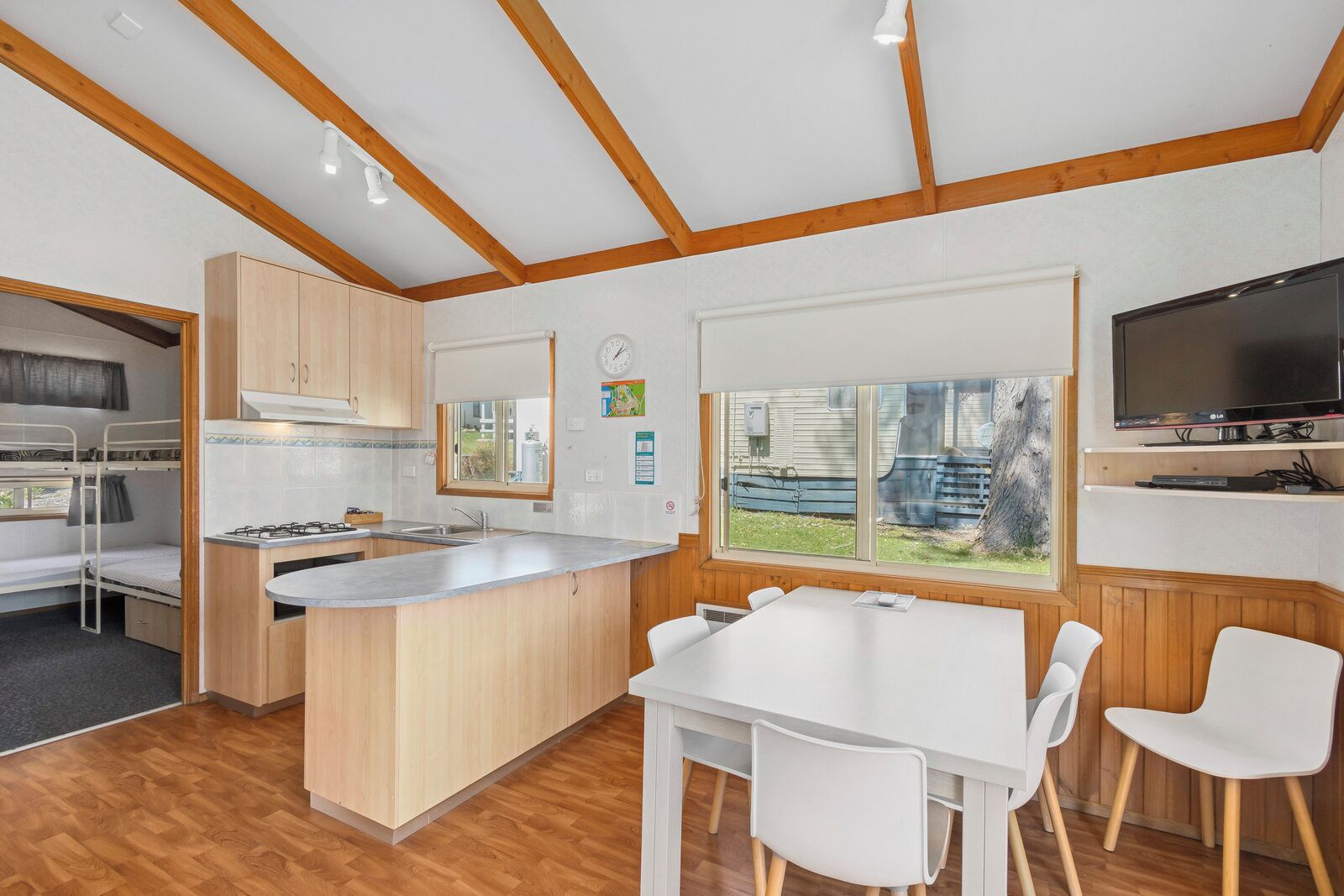 Een kleine keuken met een centraal eiland, een eethoek met witte stoelen en een aan de muur bevestigde televisie.