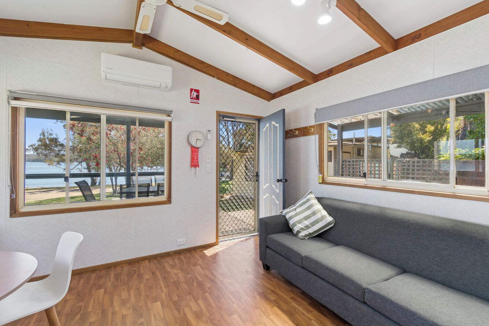 A room with a wooden ceiling, a grey sofa with a striped pillow, a white chair, and a table. There are windows with a view of trees and a building.