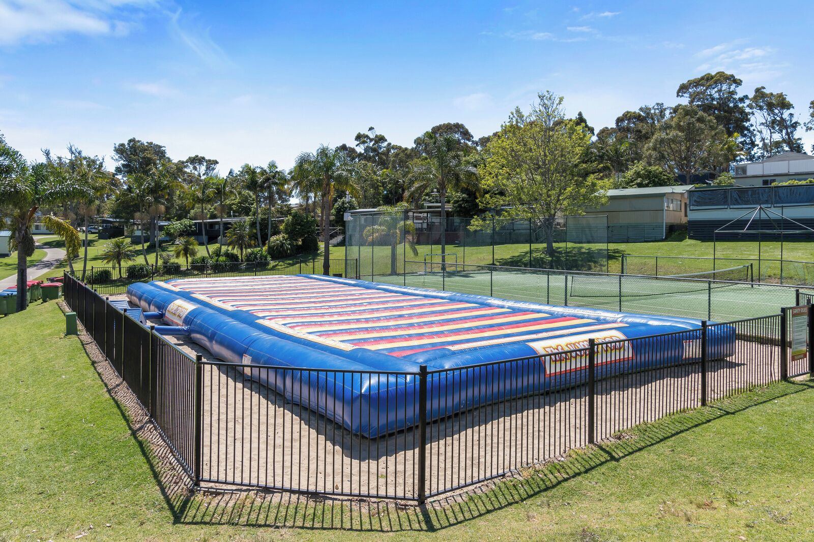 A large inflatable pool with a blue and red striped design, surrounded by a black metal fence, set in a grassy area with trees and a building in the background.