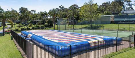 A large inflatable pool with a blue and red striped design, surrounded by a black metal fence, set in a grassy area with trees and a building in the background.