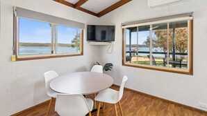 A room with a round table and four chairs, large windows with views of a lake and trees, and a mounted flat-screen TV. - Ingenia Holidays Bermagui (Wallaga Lake)