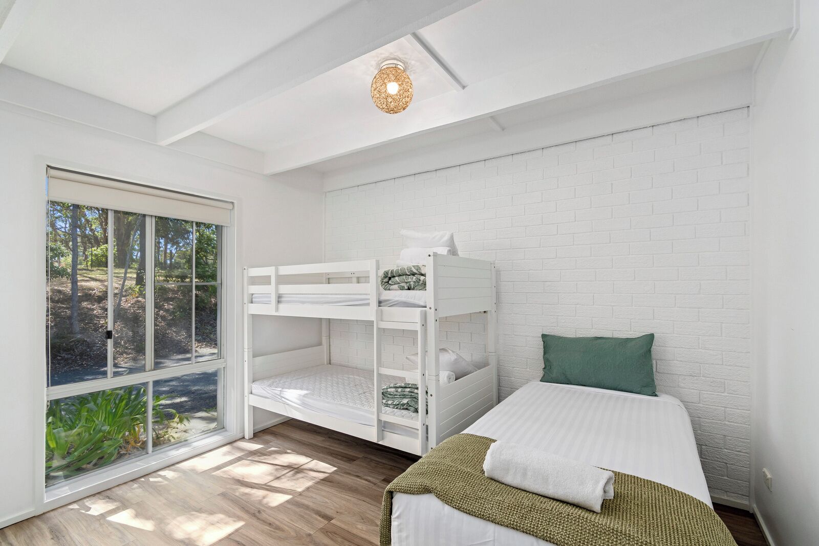 Une chambre avec un lit superposé, un lit simple, un mur en briques blanches, une grande fenêtre et un luminaire au plafond.