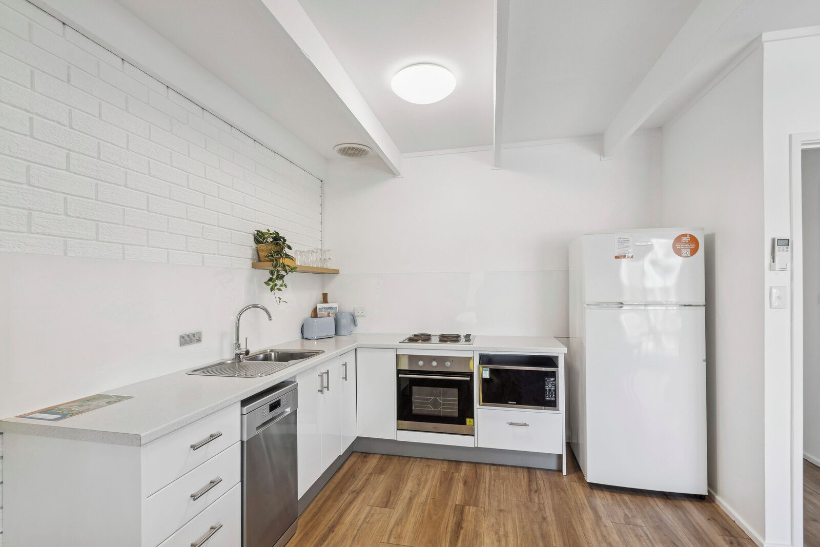 Une cuisine de taille réduite, avec des appareils électroménagers blancs, un plancher en bois et un mur en briques peintes en blanc.