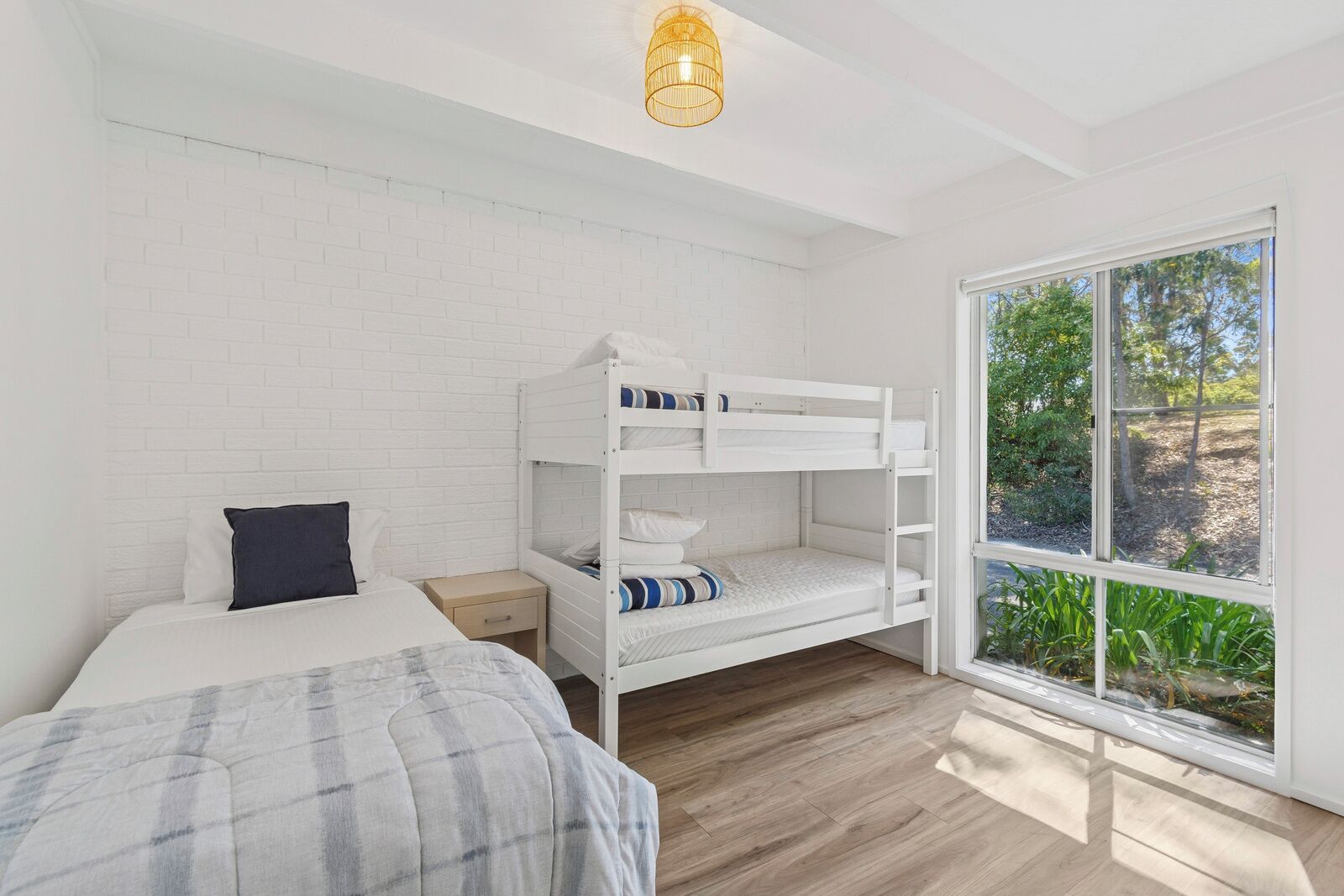 Een slaapkamer met een stapelbed, een enkel bed, een nachtkastje en een groot raam met uitzicht op groen.