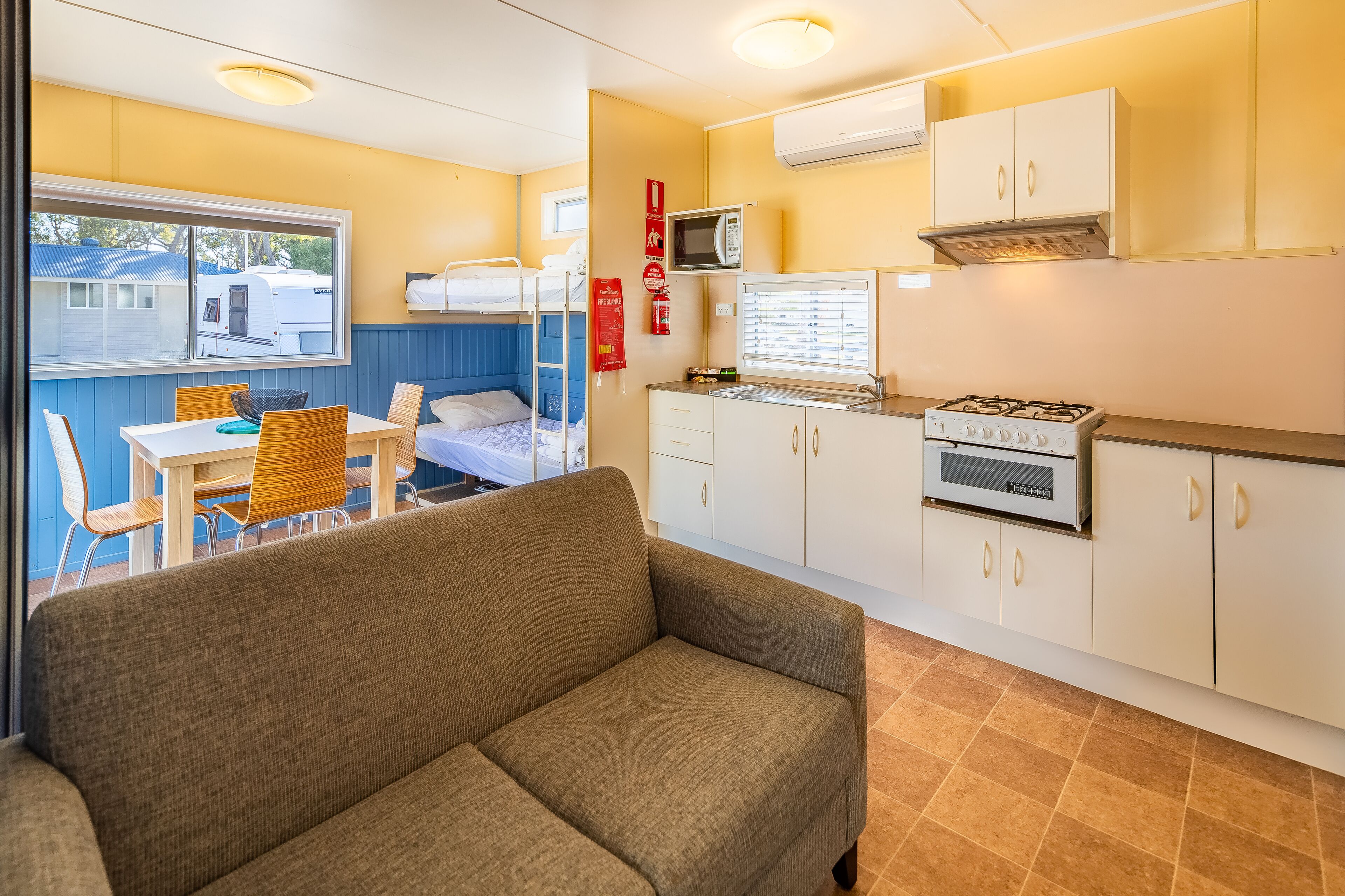 A compact kitchen with a microwave, stove, and white cabinets. There is a dining table with chairs and a sofa in the foreground.