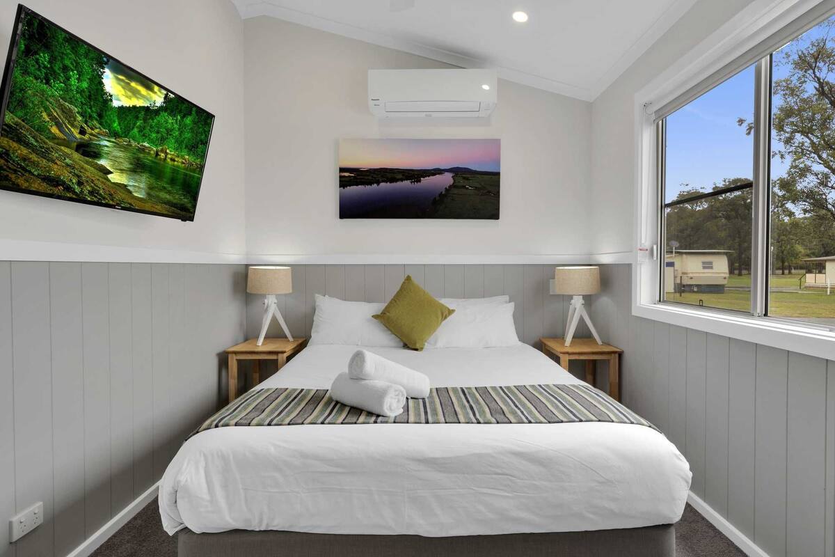A hotel room with a large bed, two bedside tables with lamps, a wall-mounted air conditioner, and a large window with a view of the outdoors.