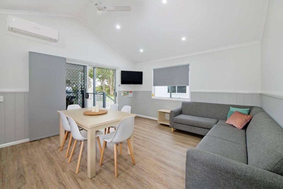 A modern living room with a grey sofa, a dining table with chairs, a flat-screen TV mounted on the wall, and a window with blinds.