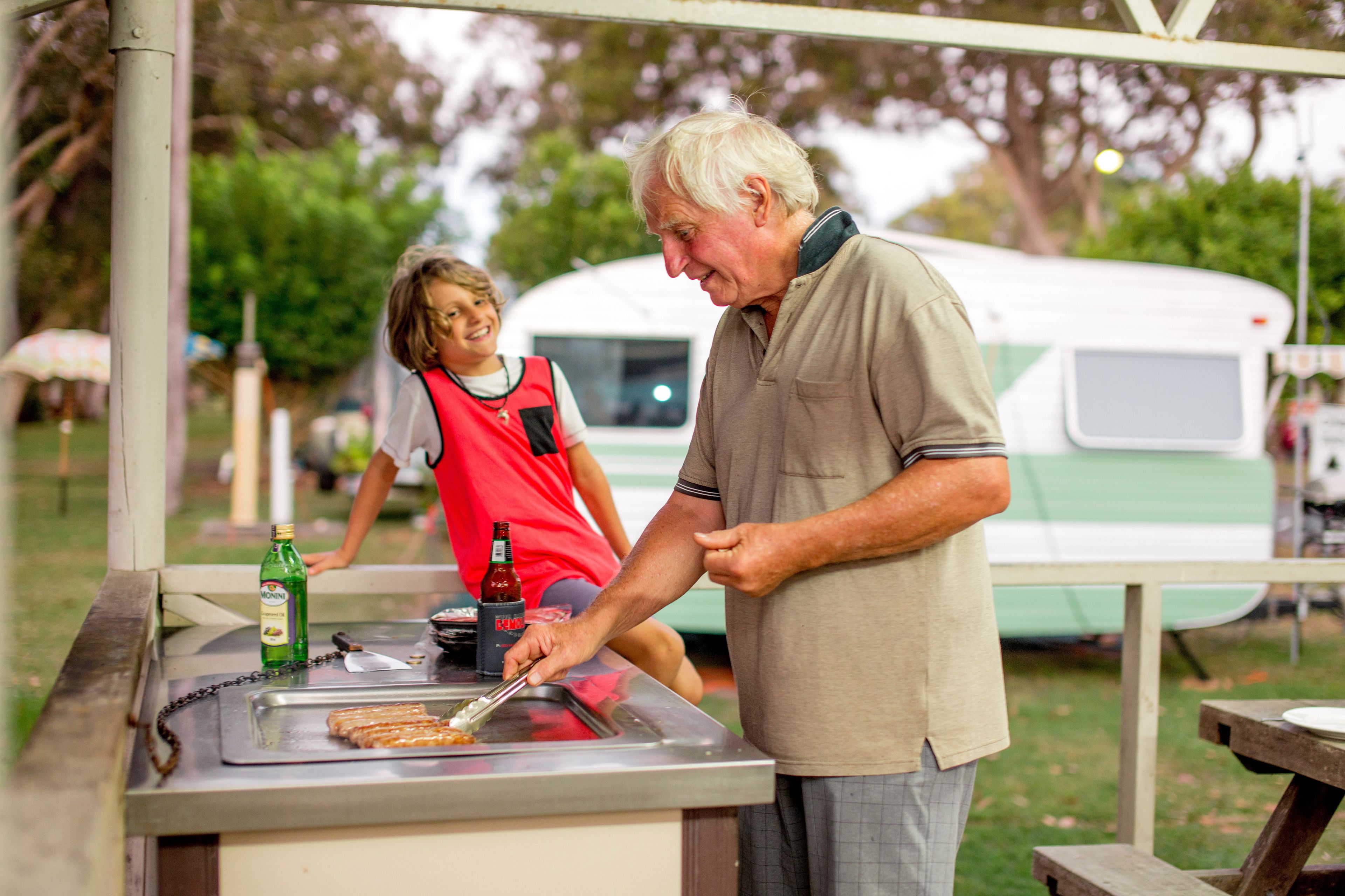 A man and a child at an outdoor food stall with a grill, a bottle of beer, and a green bottle on the counter.