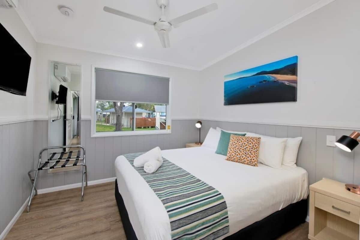 A bedroom with a bed, a nightstand, a lamp, a ceiling fan, a window, a painting, and a mirror.