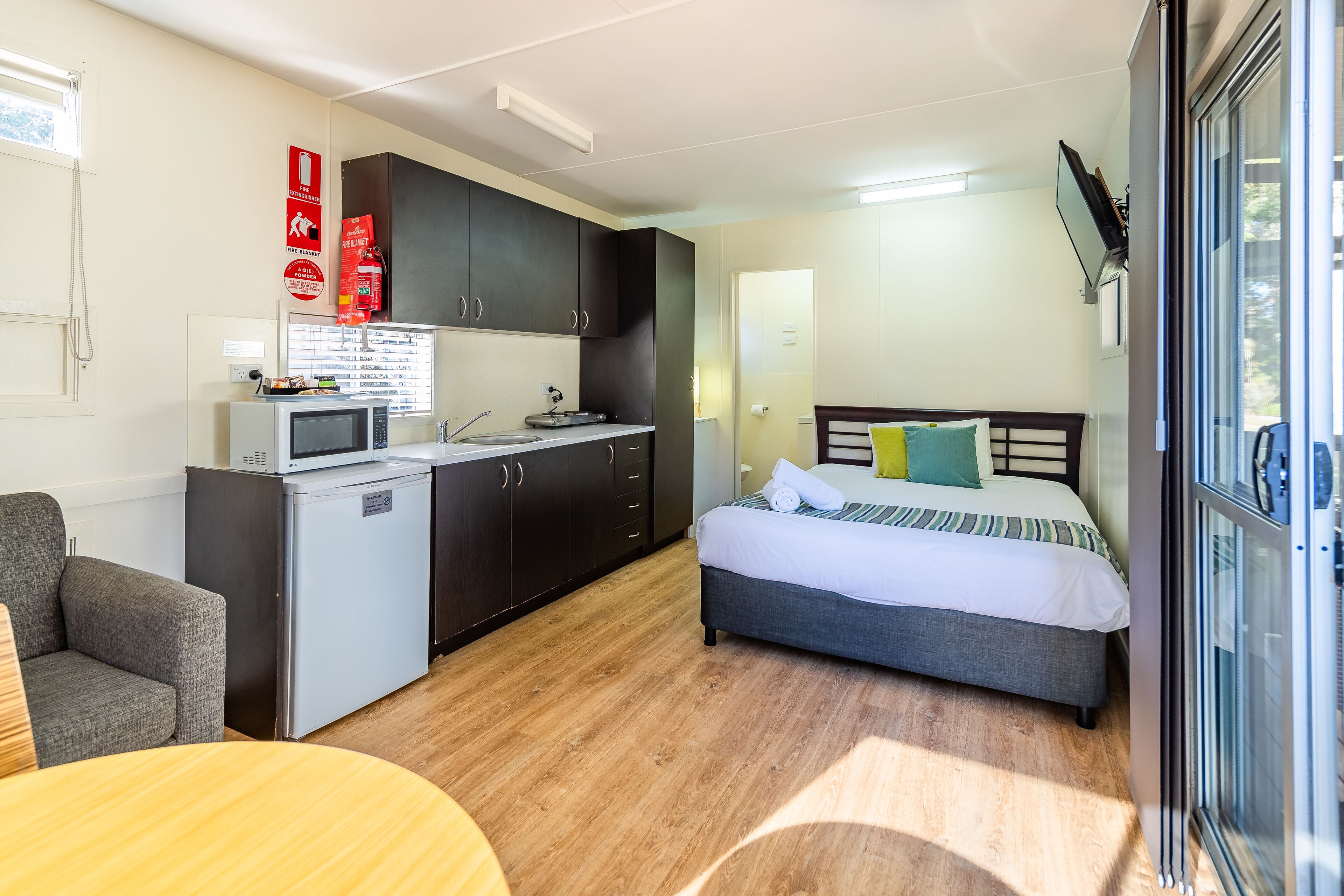A compact hotel room with a kitchenette, a bed with pillows, a sofa, and a dining table.
