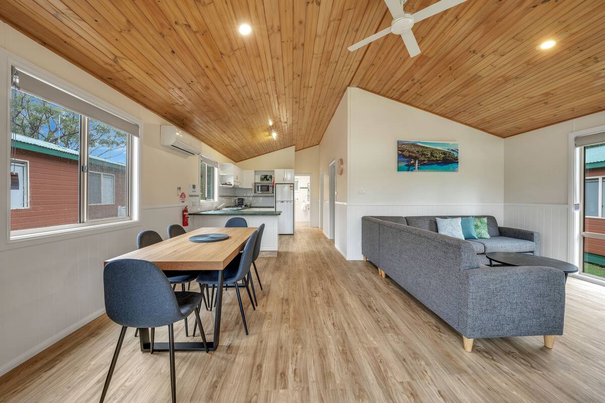 A modern kitchen with white cabinets, a dining area with a wooden table and chairs, and a grey sofa in a spacious living area with wooden flooring and a wooden ceiling.
