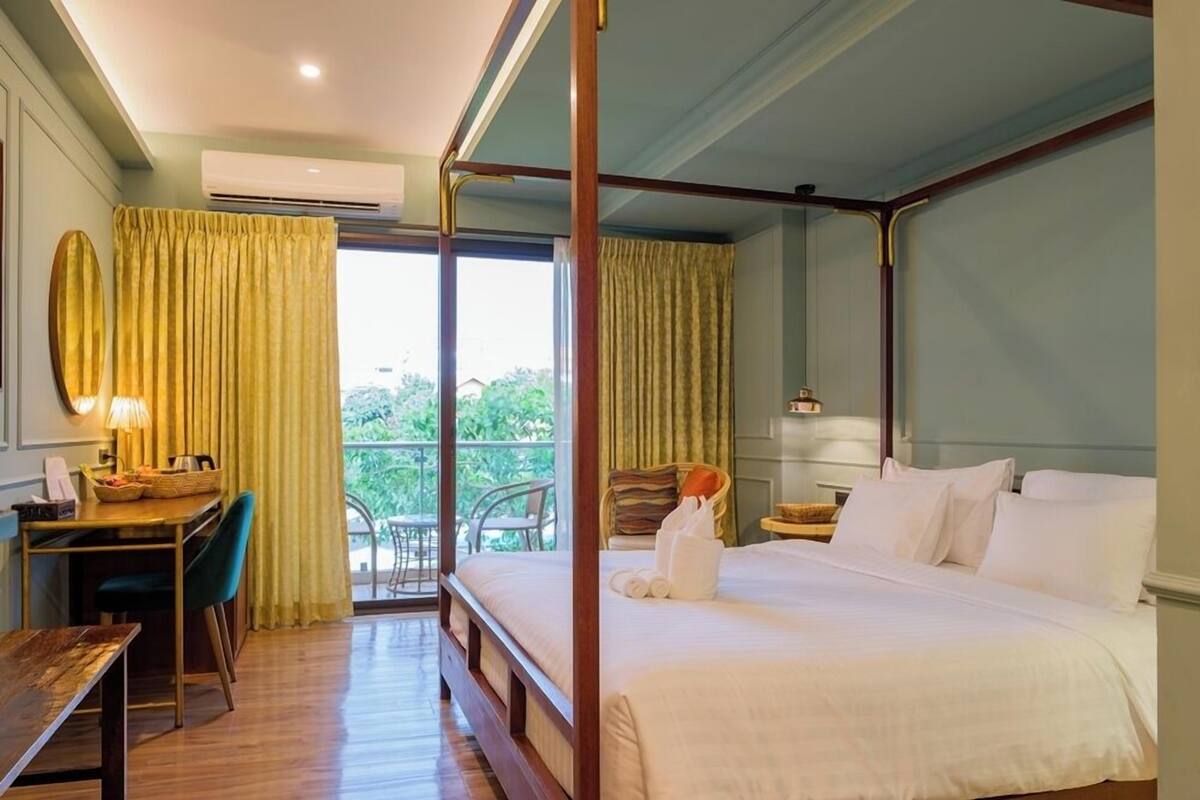 A hotel room with a four-poster bed, a desk with a chair, a small table with a lamp, and a balcony with a chair.