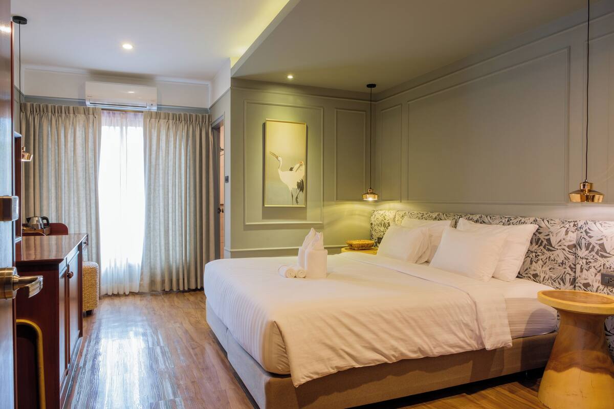 A hotel room with a large bed, a desk, and a chair. There is a window with curtains, a wall-mounted air conditioner, and a painting on the wall.