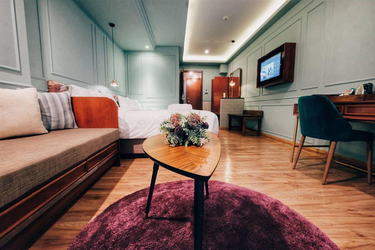 A hotel room with a bed, a wooden coffee table with flowers, a sofa, a desk, and a television on the wall.