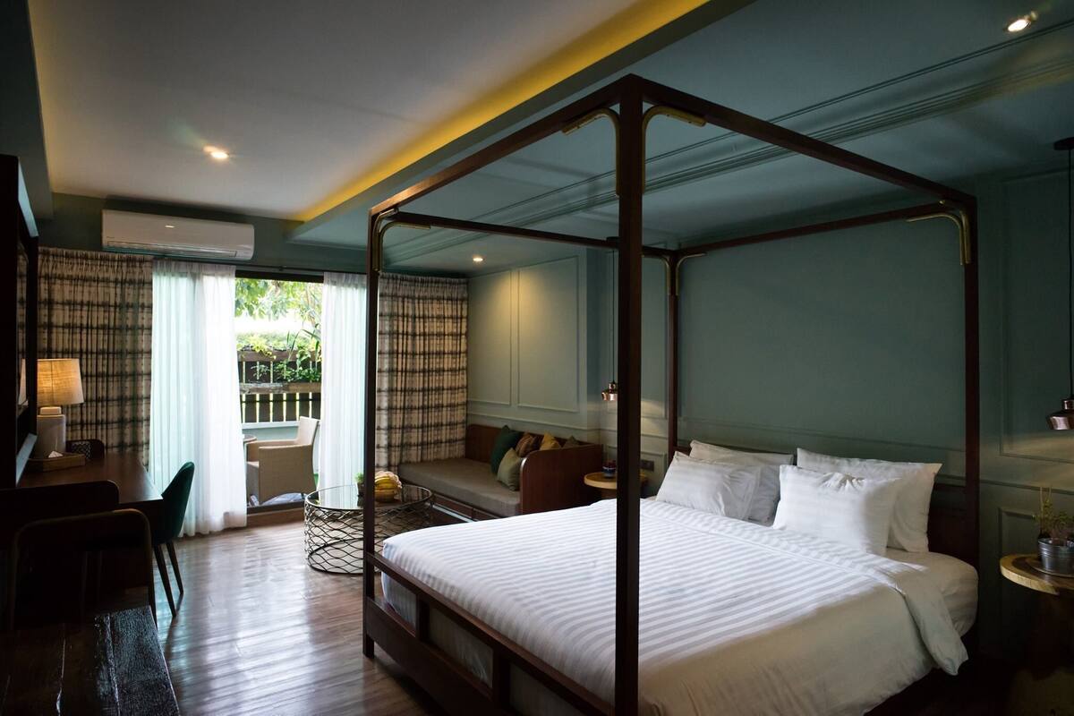 A four-poster bed with a wooden frame, a sofa, a small table, and a chair in a room with a view of greenery outside.