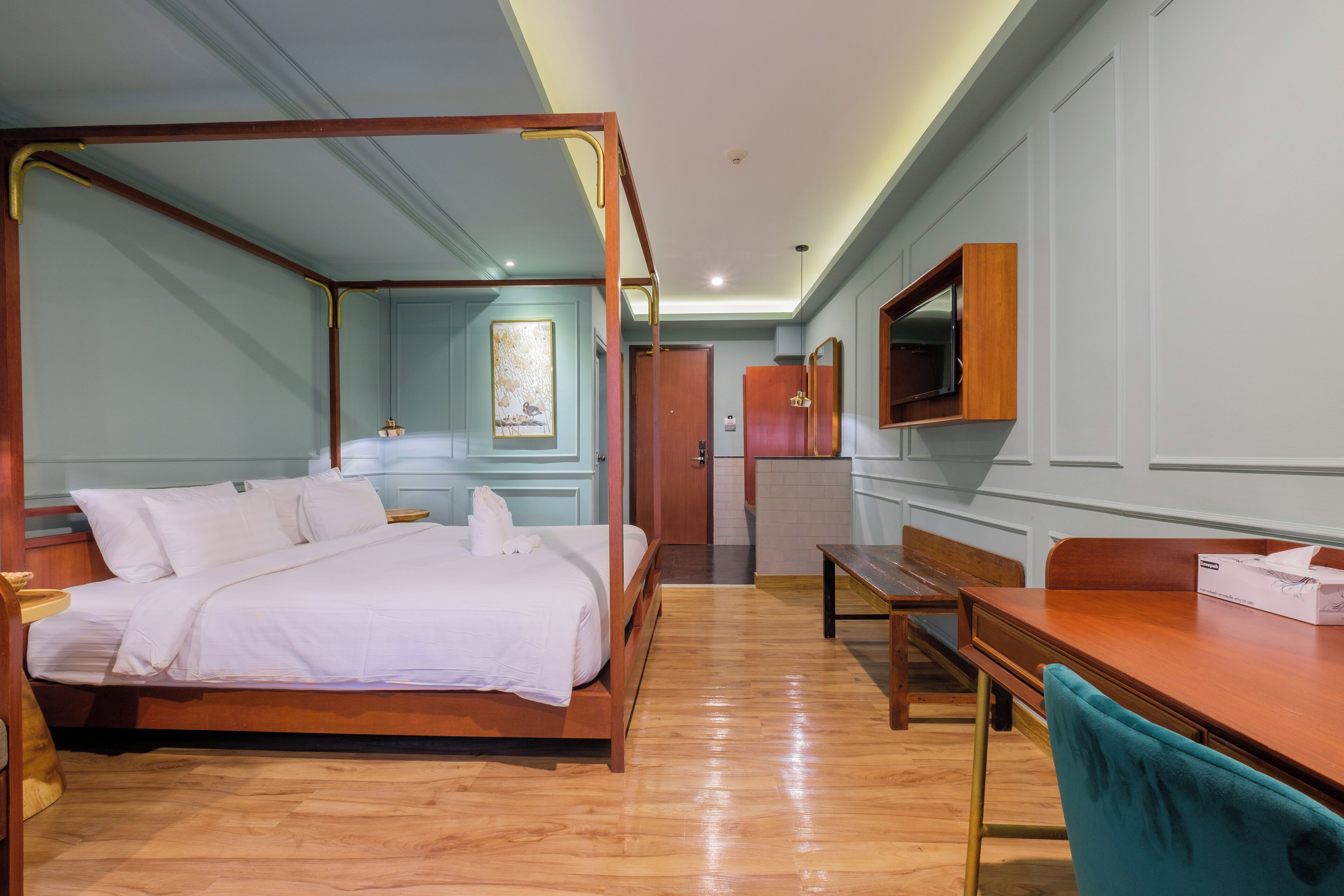 A four-poster bed with a wooden frame, a wooden desk with a chair, a television mounted on the wall, and a door leading to another room.